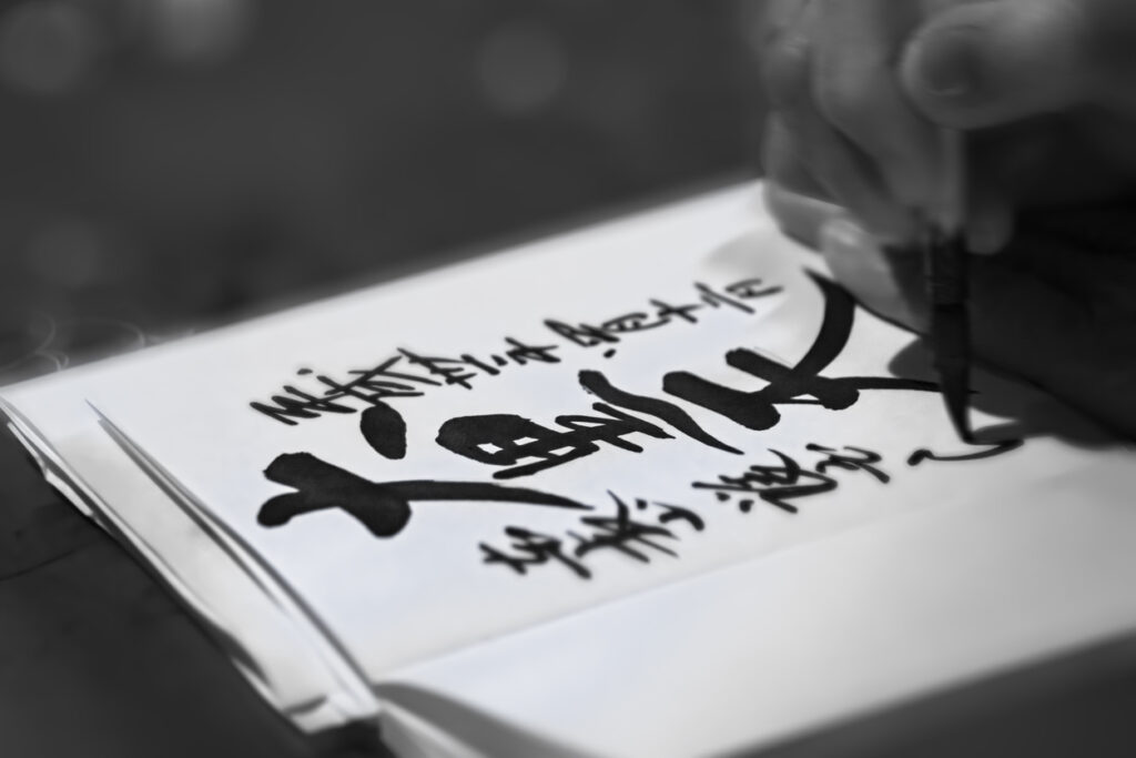 Black and white photography of a close up on a hand writing in calligraphy the Japanese name of Daikokuten the deity of wealth which is one of the seven lucky gods.