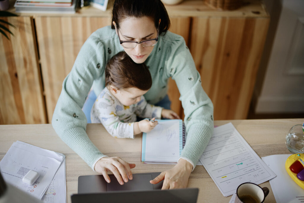 Mother holds her daughter as she takes a phone call makes a note on paper and works on her laptop in a home office