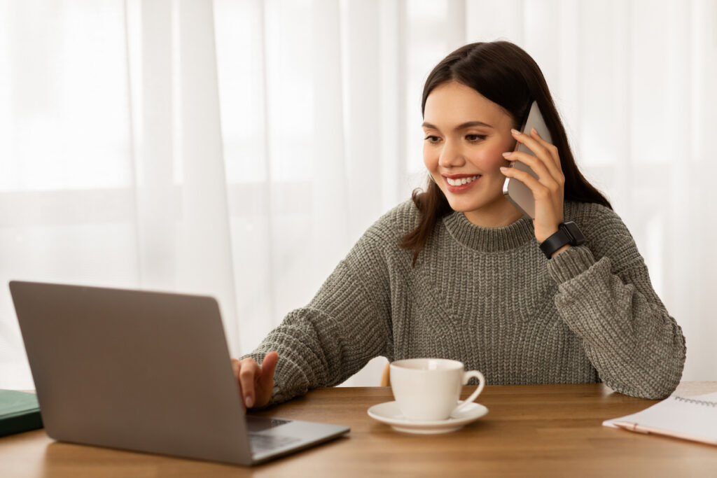 Happy young businesswoman working on laptop from home office, have phone conversation with business partner or client, drinking coffee. Entrepreneurship concept