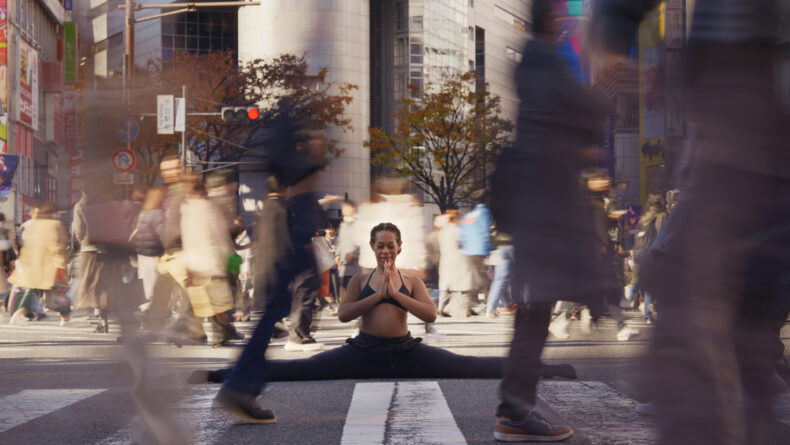 5 Meditation Tips For Life in Tokyo A woman gracefully holds a full split yoga pose in the middle of a busy crosswalk in Shibuya, Tokyo. Blurred figures of bustling pedestrians move around her, emphasizing the calm and centered stillness she exudes amidst the vibrant urban motion. Bright advertisements and iconic city buildings fill the background, adding to the dynamic energy of the scene.