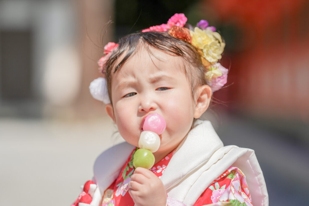 A 3-year-old Japanese girl in a red kimono celebrates Shichi-Go-San. She sits and eats dango given by her mom, who wears a pink kimono. This photo captures a sweet moment between mother and daughter.