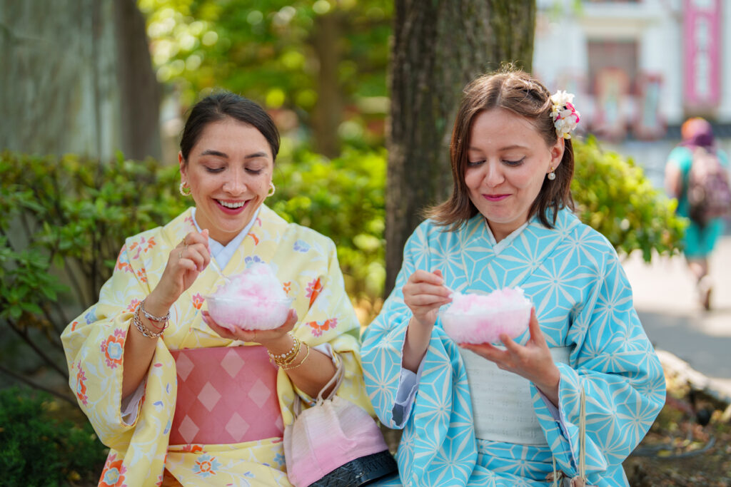 Two female tourist friends are experiencing wearing Japanese traditional clothing kimono and tasting Japanese frozen sweet food, Kamigori, in the street in Tokyo Japan.